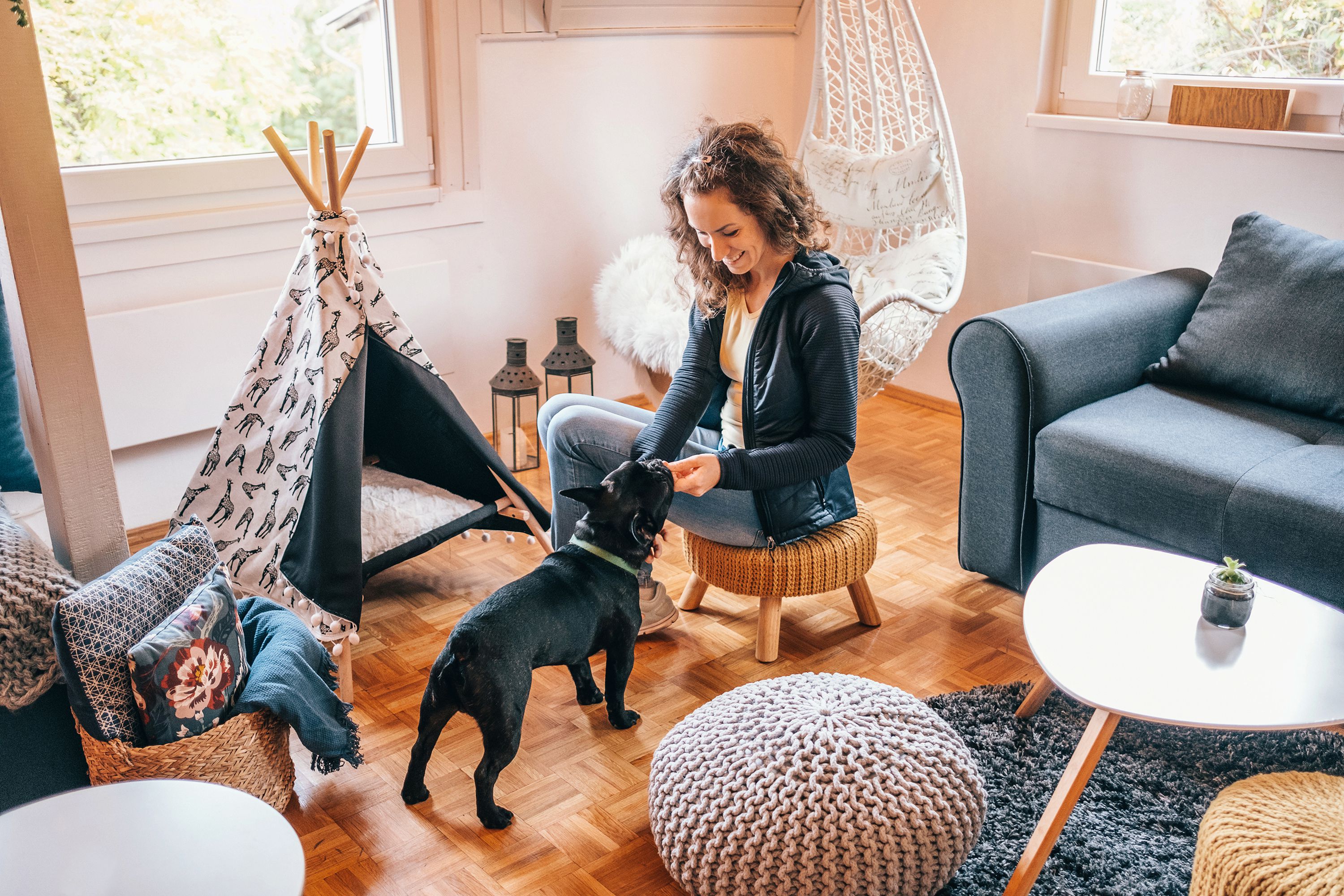 Woman sitting on a stool playing with her small black dog. They are in the living room, there is a dog teepee. 