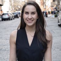 White woman with long brown hair standing on a New York City Street wearing a black sleeveless shirt.